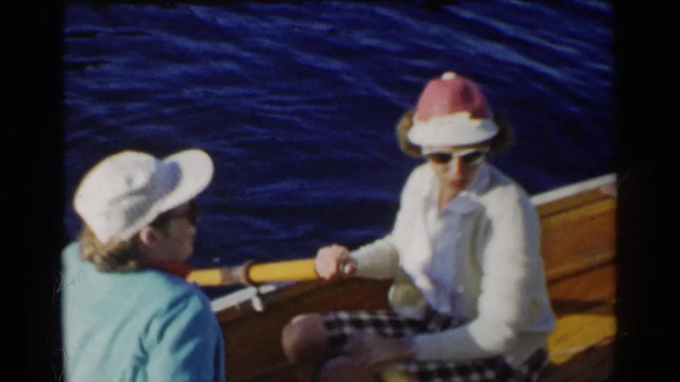 The 1955 California home-movie frame shows a woman wearing sunglasses and a red-and-white hat seated across from a companion in a white cap, with blue water behind them.