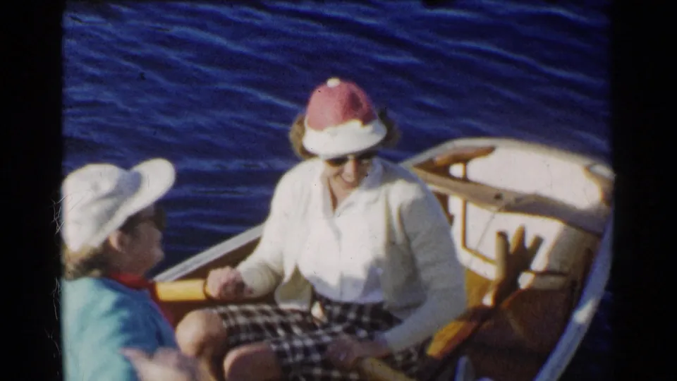 Near the end of the 1955 California clip, two older women remain seated in the small wooden boat, with the stern, paddle, hats, sunglasses, and rippling blue water visible.