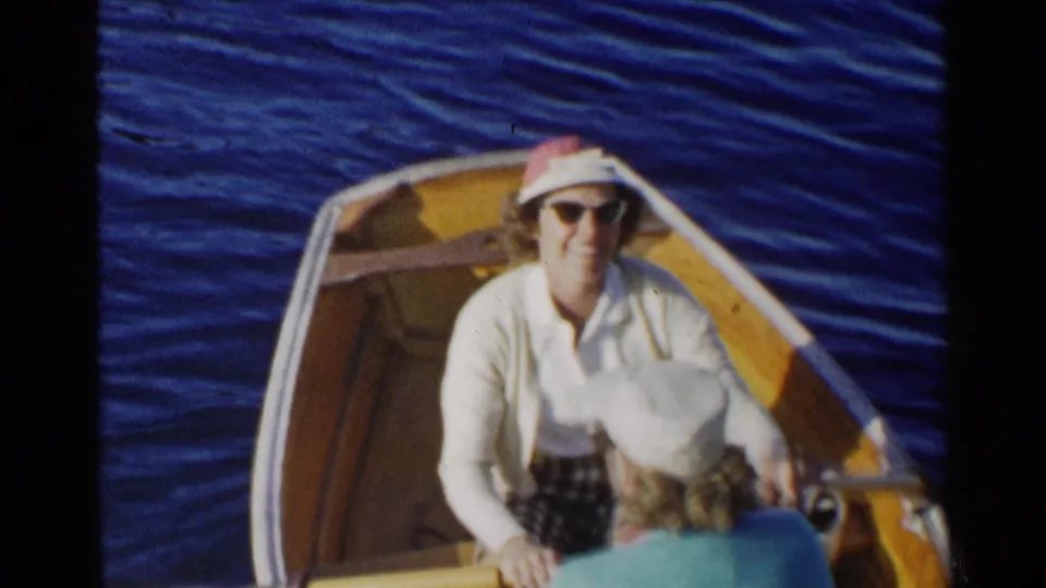 A woman in a white top, sunglasses, and red-and-white sun hat faces the camera from a small dinghy on blue water.