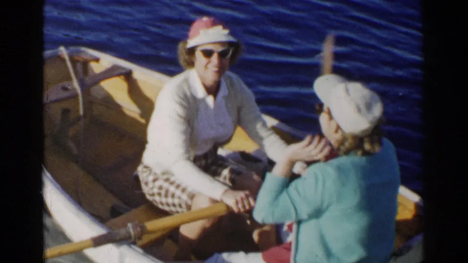 A smiling woman in sunglasses rows a small wooden dinghy as her companion in a blue sweater sits opposite her.