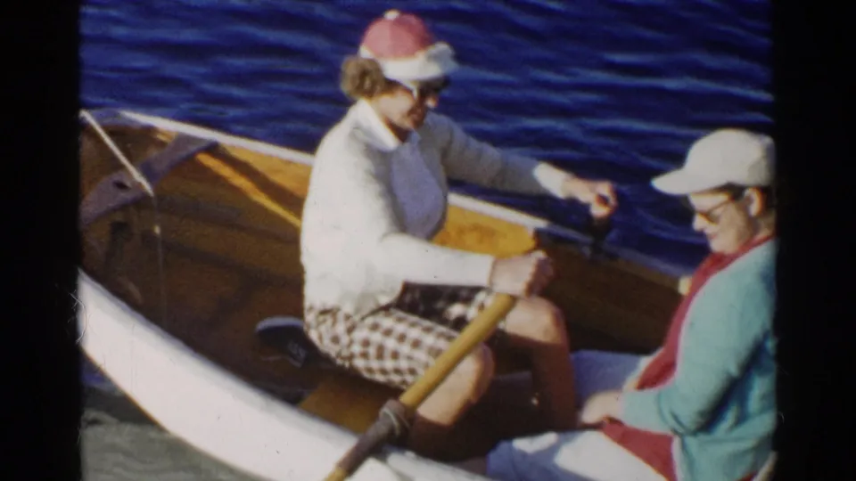 Two women share a small wooden dinghy, with one rowing and the other seated near the side against calm blue water.