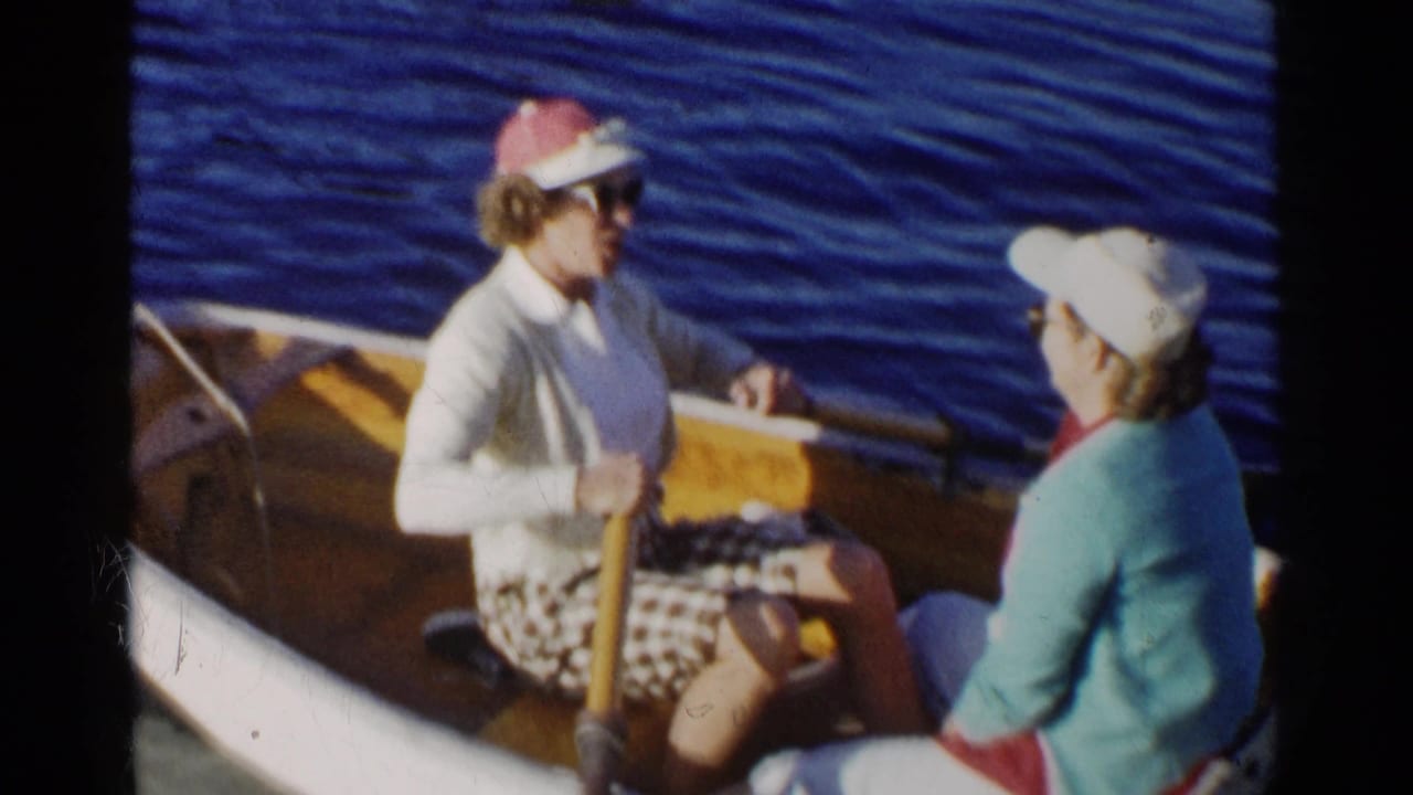 Two women in sunglasses sit in a wooden rowboat on blue lake water.