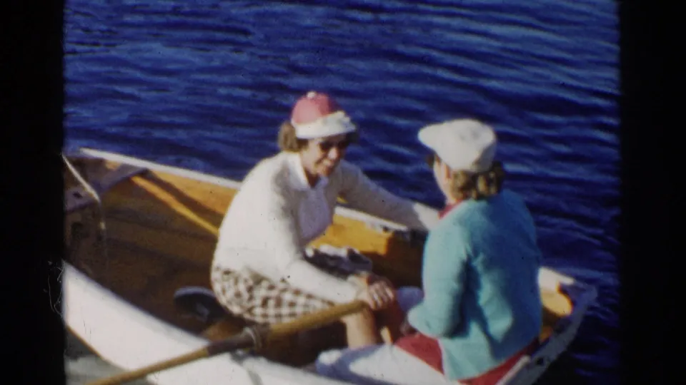 The women sit close together in a small wooden rowboat on a California lake, wearing sunglasses and light sweaters.