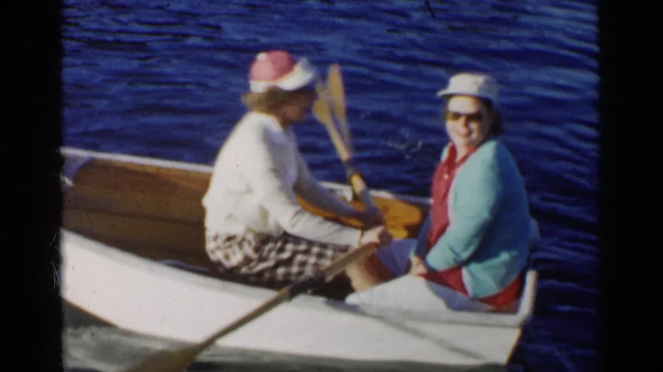 A woman in a visor holds an oar while seated opposite a friend in a turquoise cardigan in 1955 California lake footage.