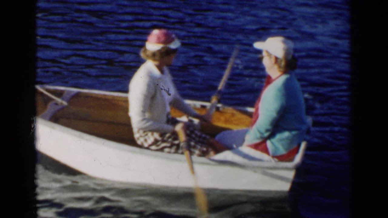 1955 California home-movie frame of two women in hats and sweaters seated in a small white rowboat on a lake.