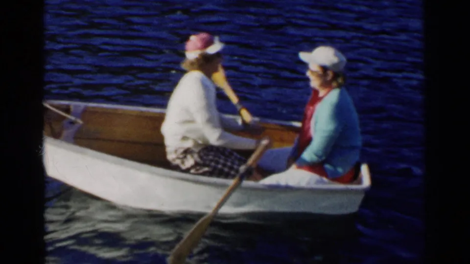 Two women ride in a white rowboat on a California lake in 1955, with one rowing and the other seated in a blue sweater.