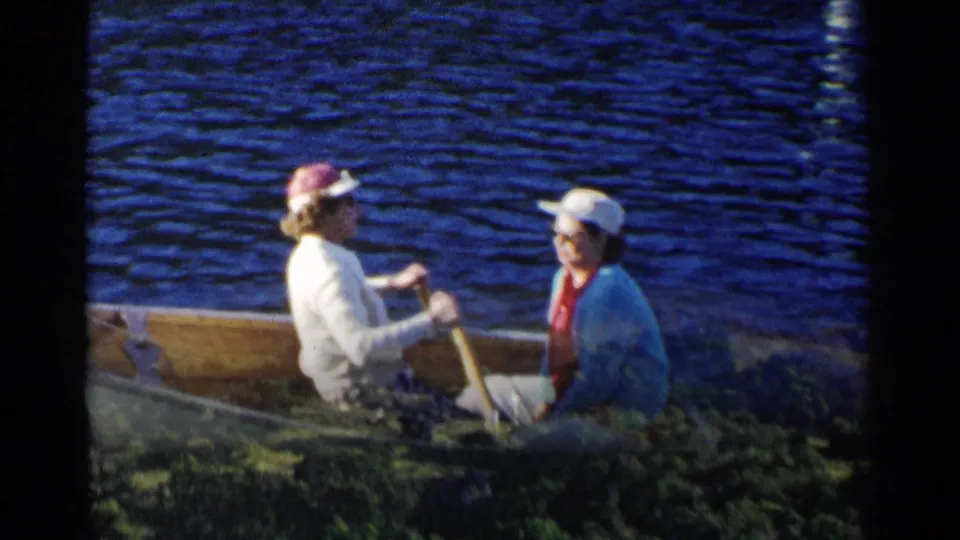A 1955 California home-movie frame shows two women in a rowboat near the water’s edge, partly framed by foreground vegetation.