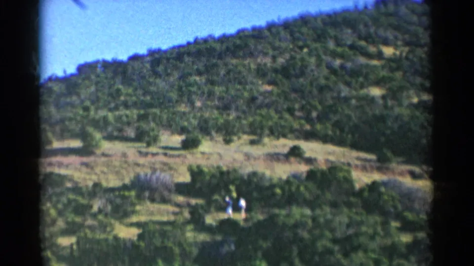 In this 1955 California 8mm frame, two small figures in light clothing walk through a dry, tree-covered hillside landscape.