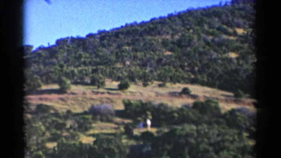 Two distant people are visible near the lower part of a sunlit California hillside, framed by trees, brush, and 8mm film borders.