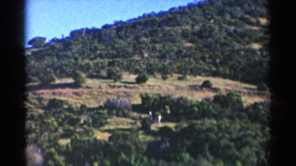Two distant figures in light clothing appear near brush at the base of a tree-covered California hill in 1955 home-movie footage.