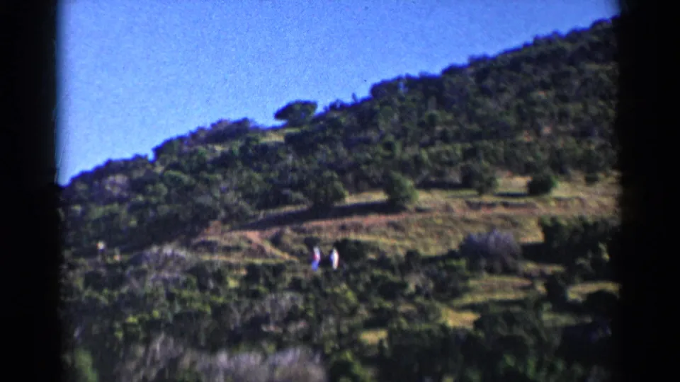 1955 California home-movie frame showing two distant walkers against a green hillside and bright blue sky.