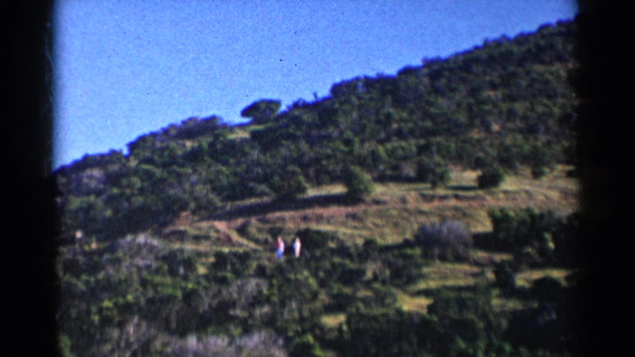 1955 California home-movie frame showing green hills, blue sky, scrub, and two distant walkers on a dirt path.
