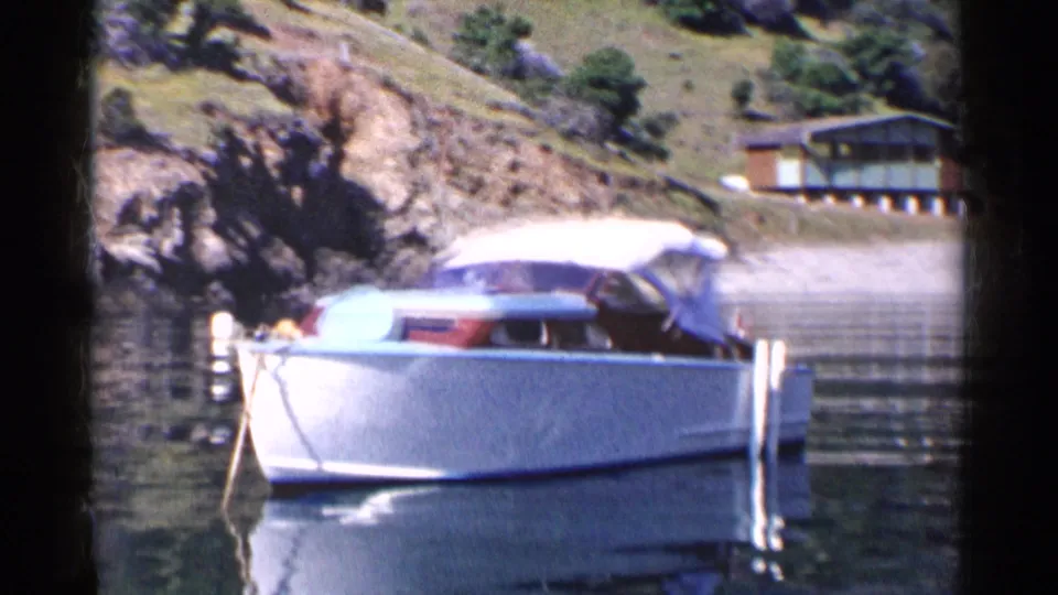 1955 California waterfront frame showing a white motorboat with canopy, calm water, rocky shore, and a cabin behind it.