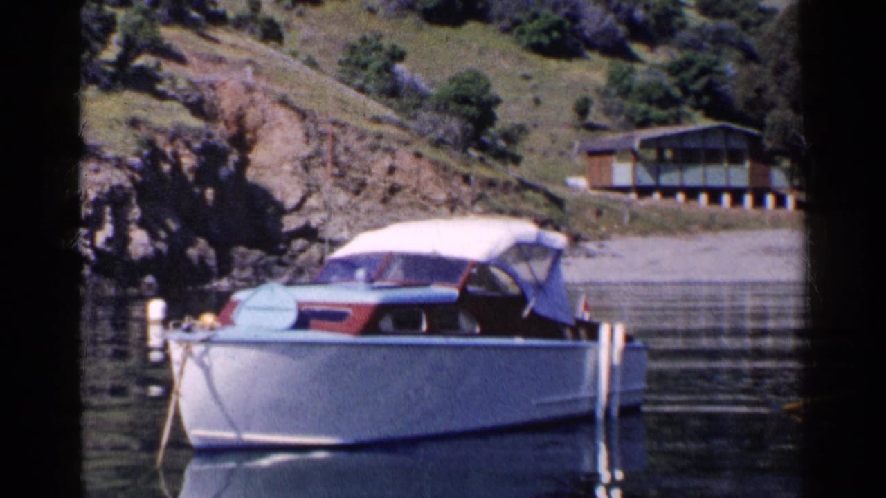 1955 California home-movie frame of a white motorboat with red and blue trim moored near a rocky shore and hillside house.