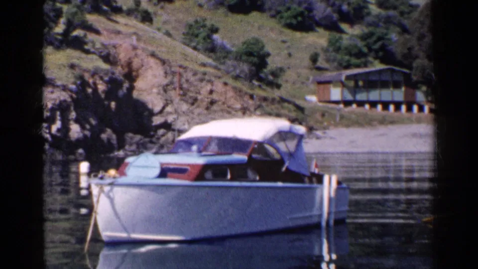 1955 California home-movie frame of a white motorboat with red and blue trim moored near a rocky shore and hillside house.