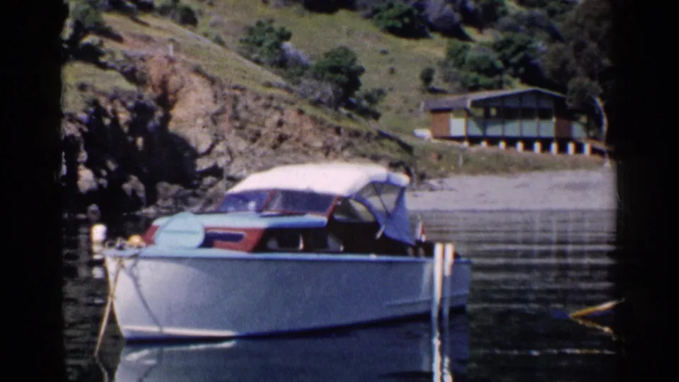 1955 California frame showing a moored white motorboat on calm water, with a rocky bank and modern-looking house in the background.