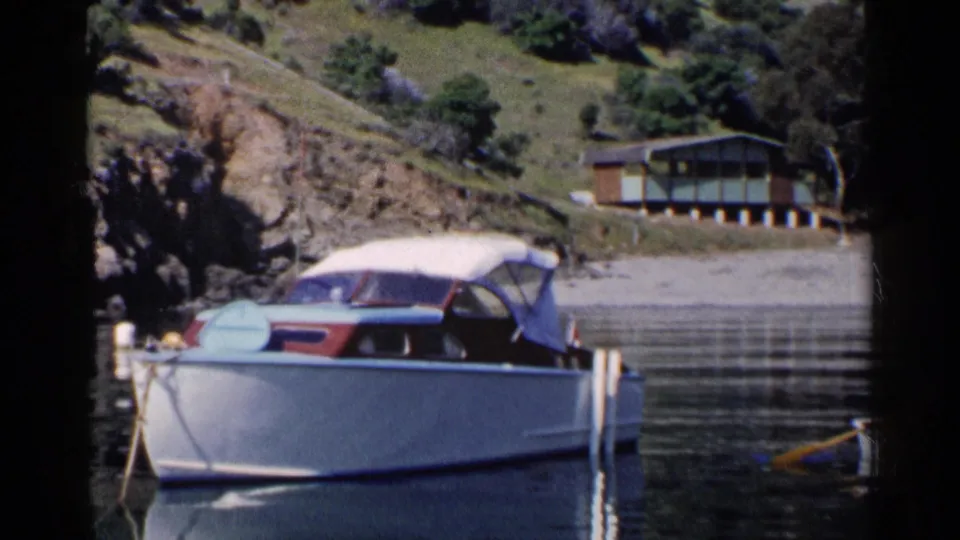 1955 California home-movie frame of a classic moored motorboat on still water beneath a hillside house and rocky shore.