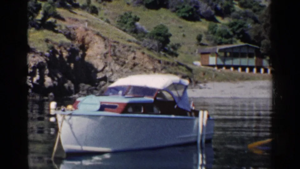 1955 California frame of a white motorboat with canvas top, moored on calm water near a rocky shoreline and hillside house.