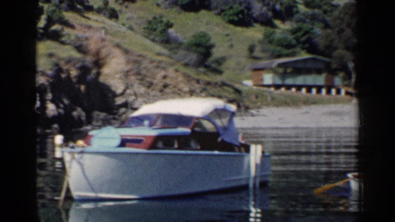 Docked canopy motorboat beside calm lake water with hillside cabins in the background.