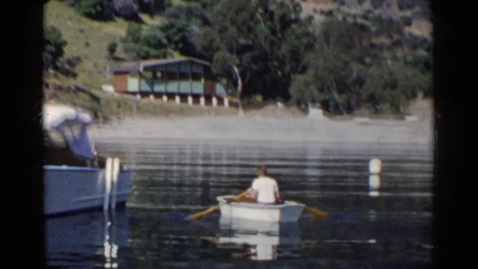 1955 California lake scene with a man in a white shirt rowing a small boat near a docked canopy motorboat and shoreline cabin.