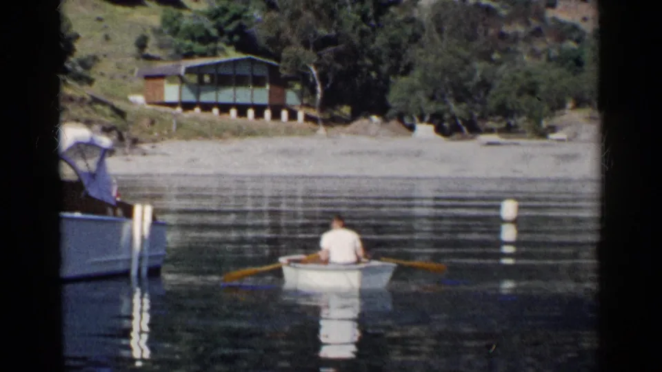 A man rows a small boat away from camera on a California lake, with a canopy motorboat at frame edge and a cabin on the opposite shore.