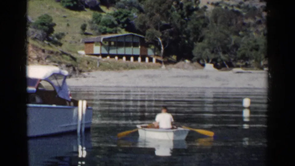 1955 California home-movie frame of a man rowing across still lake water below a small lakeside cabin and wooded hillside.