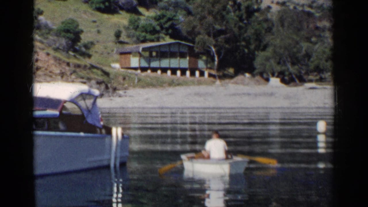 Man rows a small white boat on calm lake water near a larger moored boat and hillside cabin.