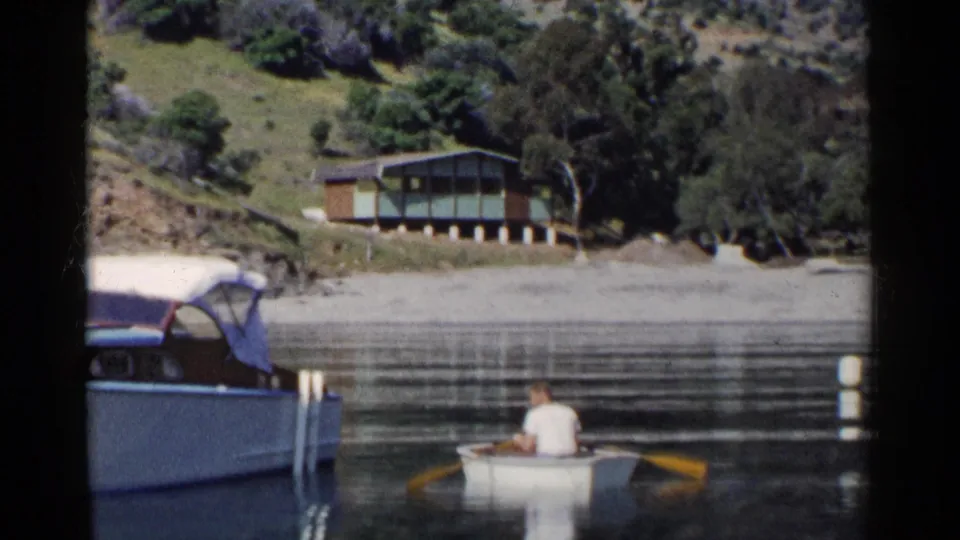 1955 California footage frame with a man in a white rowboat beside calm water, shoreline buoys, a moored boat, and a cabin beyond.