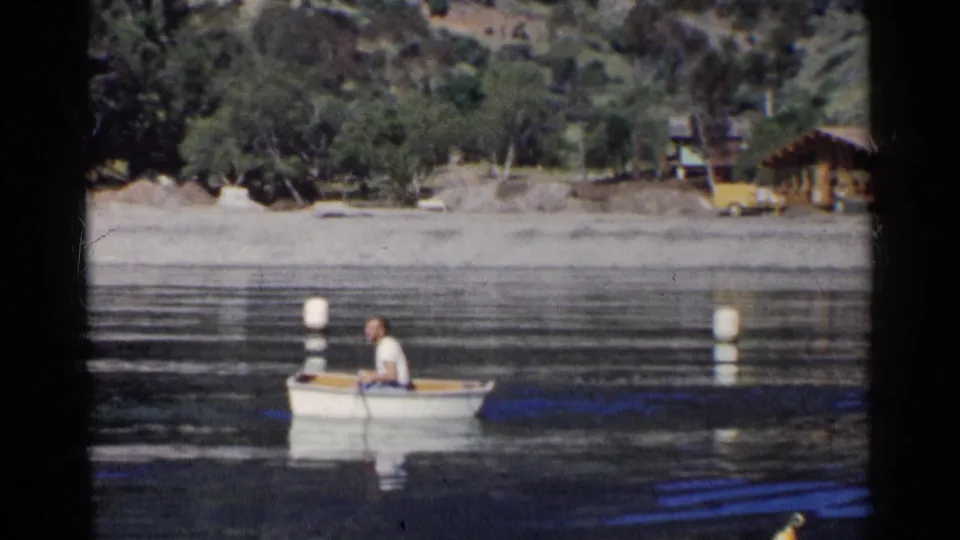 1955 California home-movie frame showing a man rowing a white boat across reflective lake water below a wooded shoreline and small cabins.