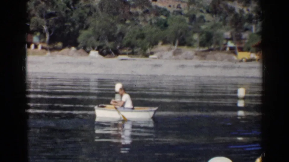 1955 California frame showing a capped man rowing a small white boat on calm water, with buoys and a wooded lakeshore behind him.