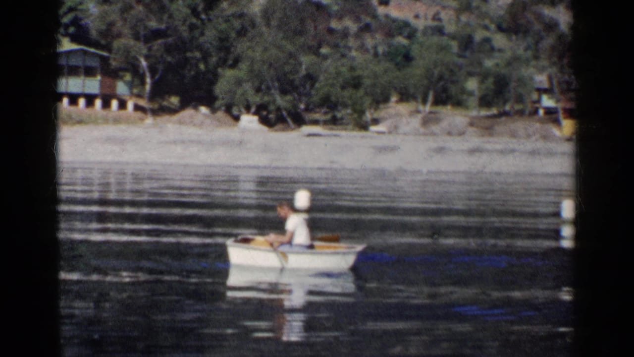 Man rows a small boat on calm water with a lakeside cabin and wooded hillside behind him.