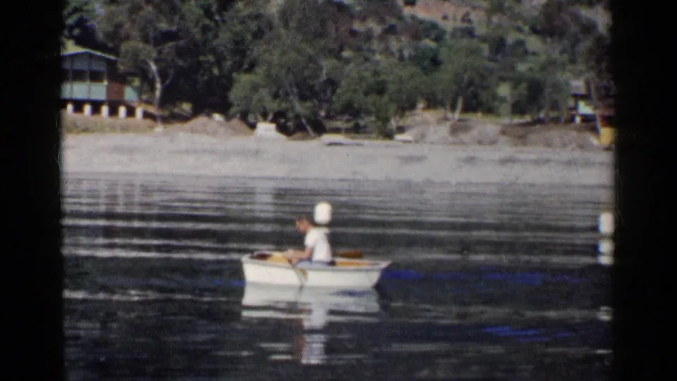 1955 California 8mm frame of a man in a white shirt rowing a small boat near a shoreline cabin and wooded hillside.