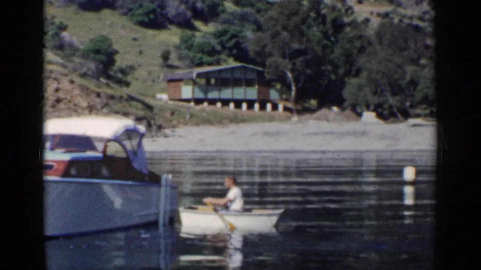 1955 California home-movie frame showing a man in a rowboat beside a larger motorboat, with a lakeside cabin in the background.