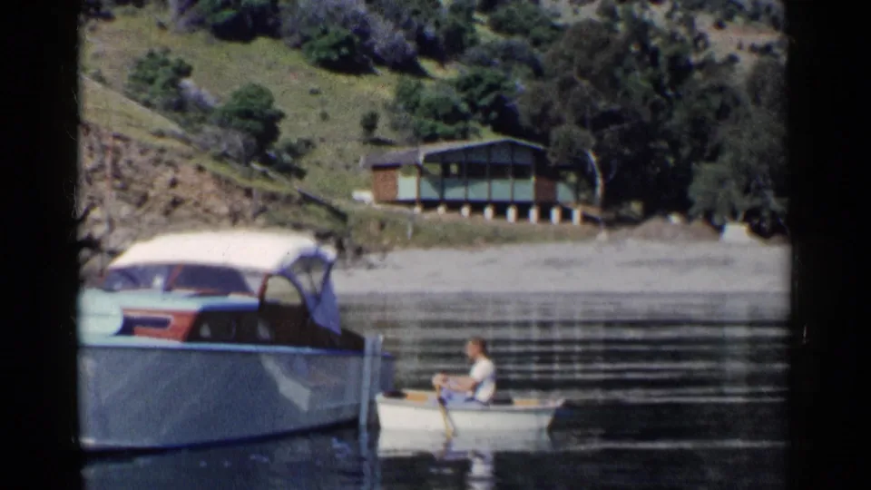 1955 California 8mm still of a man in a small rowboat next to a larger boat, with reflective water and a hillside cabin beyond.