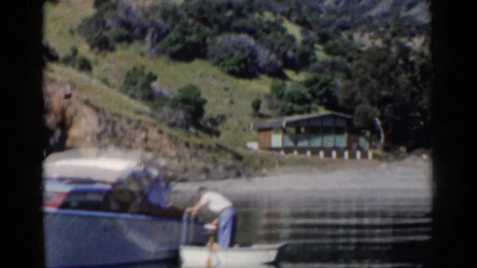 1955 California home-movie frame of a man leaning from a rowboat beside a larger boat, with shoreline trees and a cabin behind.