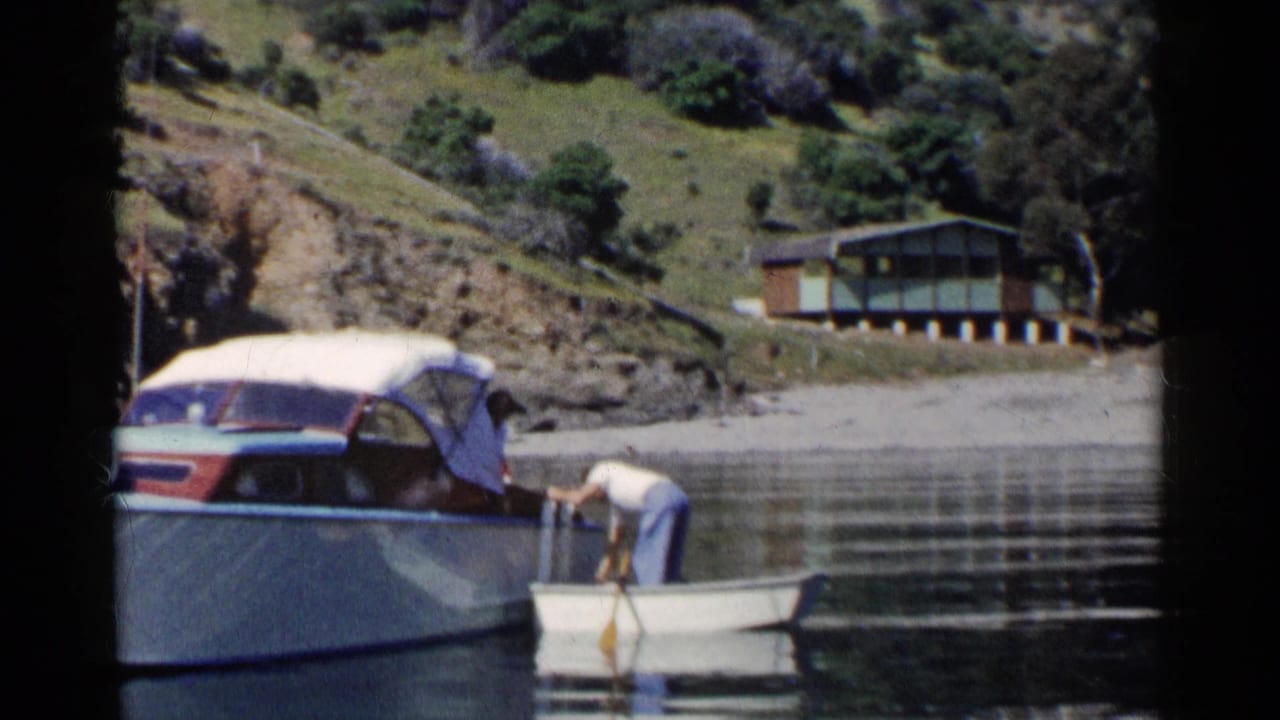 Man bends over a small boat beside a larger cabin cruiser on a calm lake.
