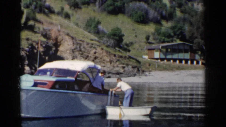 The man steadies himself near the small boat and cabin cruiser, with reflective lake water and a hillside house in the background.