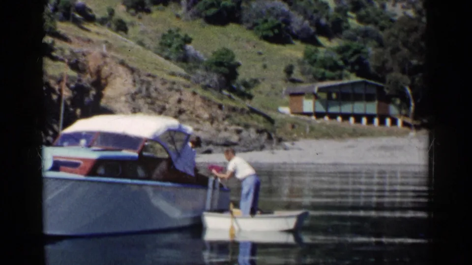 A man in casual clothing reaches toward the cabin cruiser from the dinghy during a quiet California lakeside boating moment.
