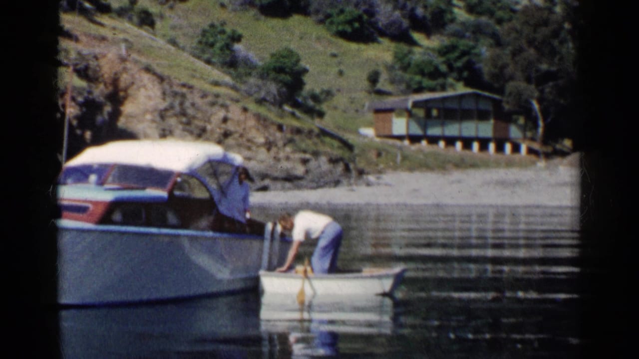 Man in a rowboat reaches near a covered motorboat on a calm lake.