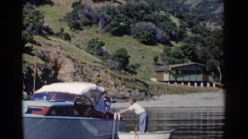 A woman in a covered motorboat leans toward a man standing in a rowboat on a California lake in 1955.