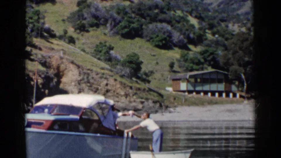 The rowboat drifts beside the covered motorboat while the man extends an arm across the water in 1955 California footage.