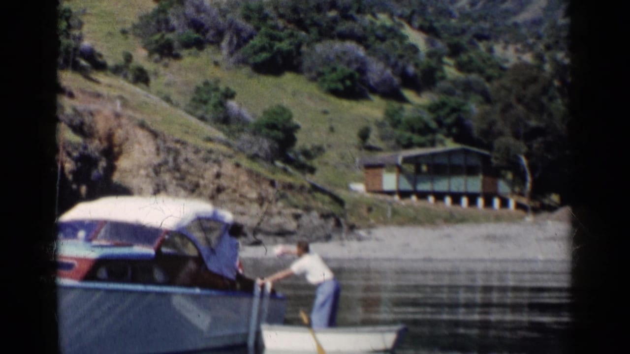 1955 California home-movie frame showing a man rowing a small boat alongside a larger covered motorboat near green hills.