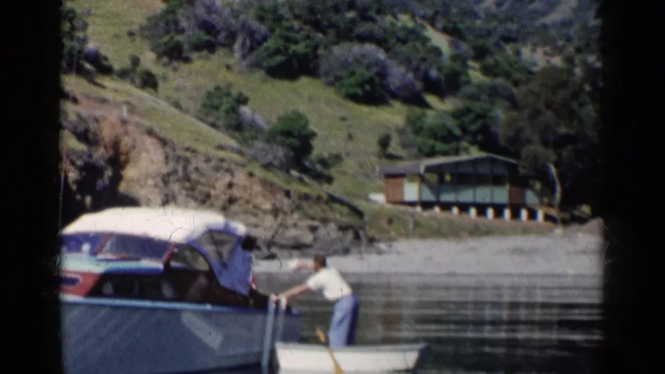 1955 California home-movie frame showing a man rowing a small boat alongside a larger covered motorboat near green hills.