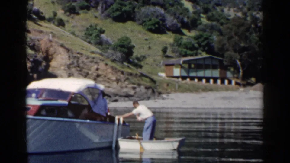 A man in a small rowboat maneuvers beside a larger motorboat on calm California lake water in 1955 footage.