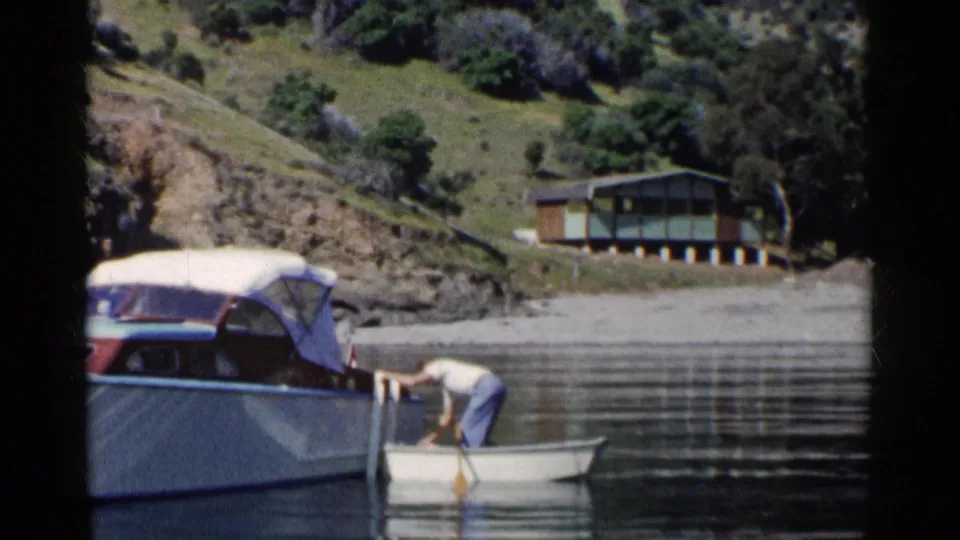 1955 California lakeside footage showing a man leaning in a rowboat beside a covered motorboat with a cabin on shore.