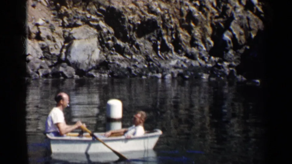 A man and another passenger sit in a rowboat on calm California lake water, with a rocky shore behind them.