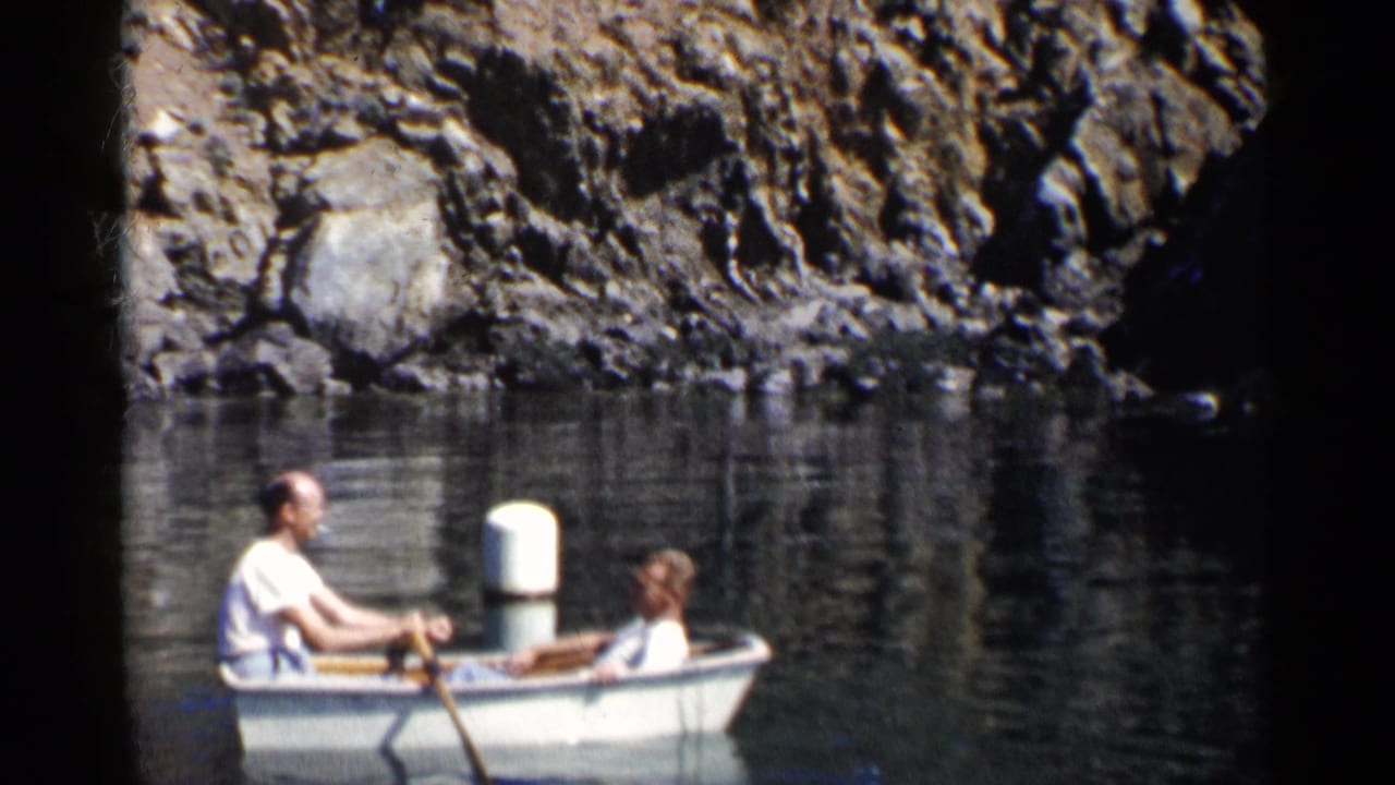 1955 California 8mm frame showing two men in a small white rowboat near dark rocky shoreline and still water.