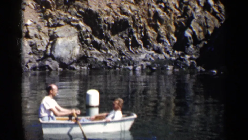 1955 California 8mm frame showing two men in a small white rowboat near dark rocky shoreline and still water.