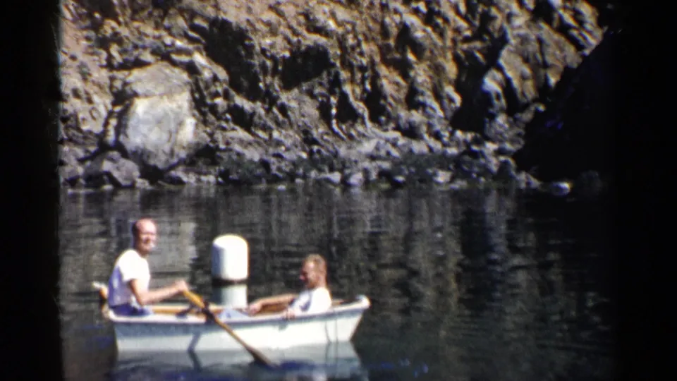 1955 California home-movie frame with one man rowing and another seated in a white boat beside rocky water.