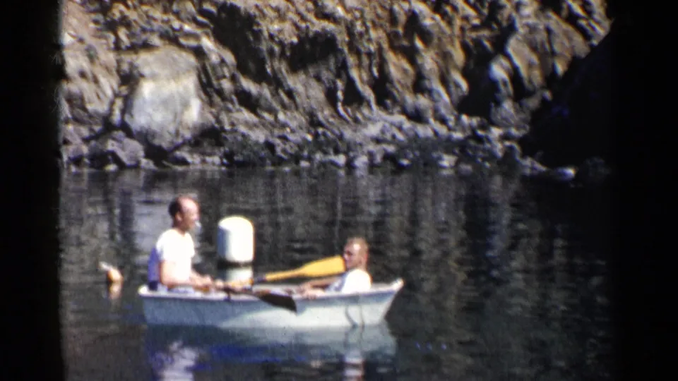Two men in a white rowboat move across calm California water, with a dark rock face behind them in this 1955 8mm frame.